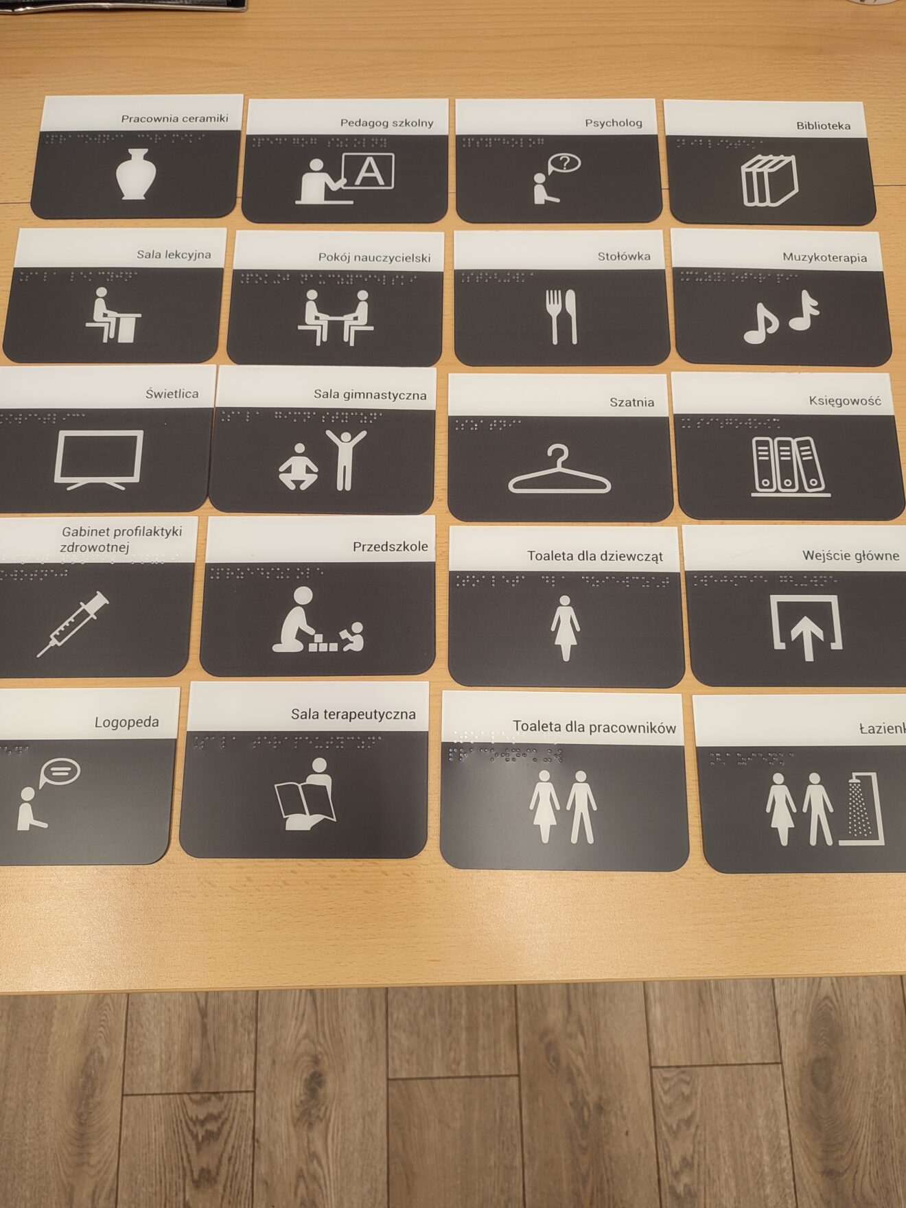 Czarnobiałe tabliczki z nazwami pomieszczeń szkolnych, alfabetem Braille’a i piktogramami