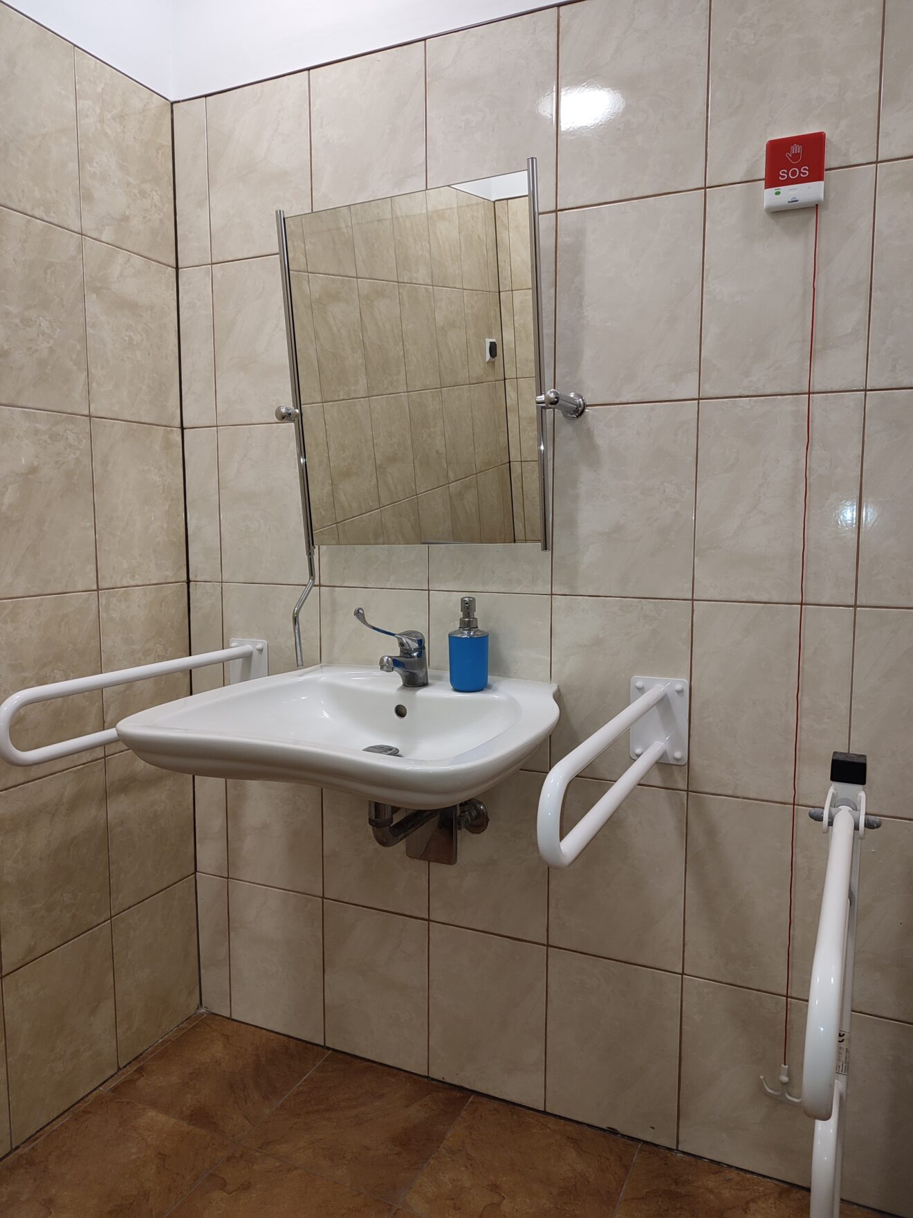 Pomieszczenie toalety. Na ścianie zawieszone lustro, umywalka z uchwytami i czerwony przycisk alarmowy
