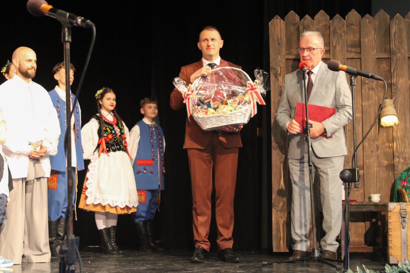 Na scenie przedstawiciele Powiatu Tomaszowskiego jeden z nich trzyma kosz upominkowy, drugi przemawia do mikrofonu. W tle dzieci w strojach ludowych z Zespołu Folklorystycznego Roztocze.