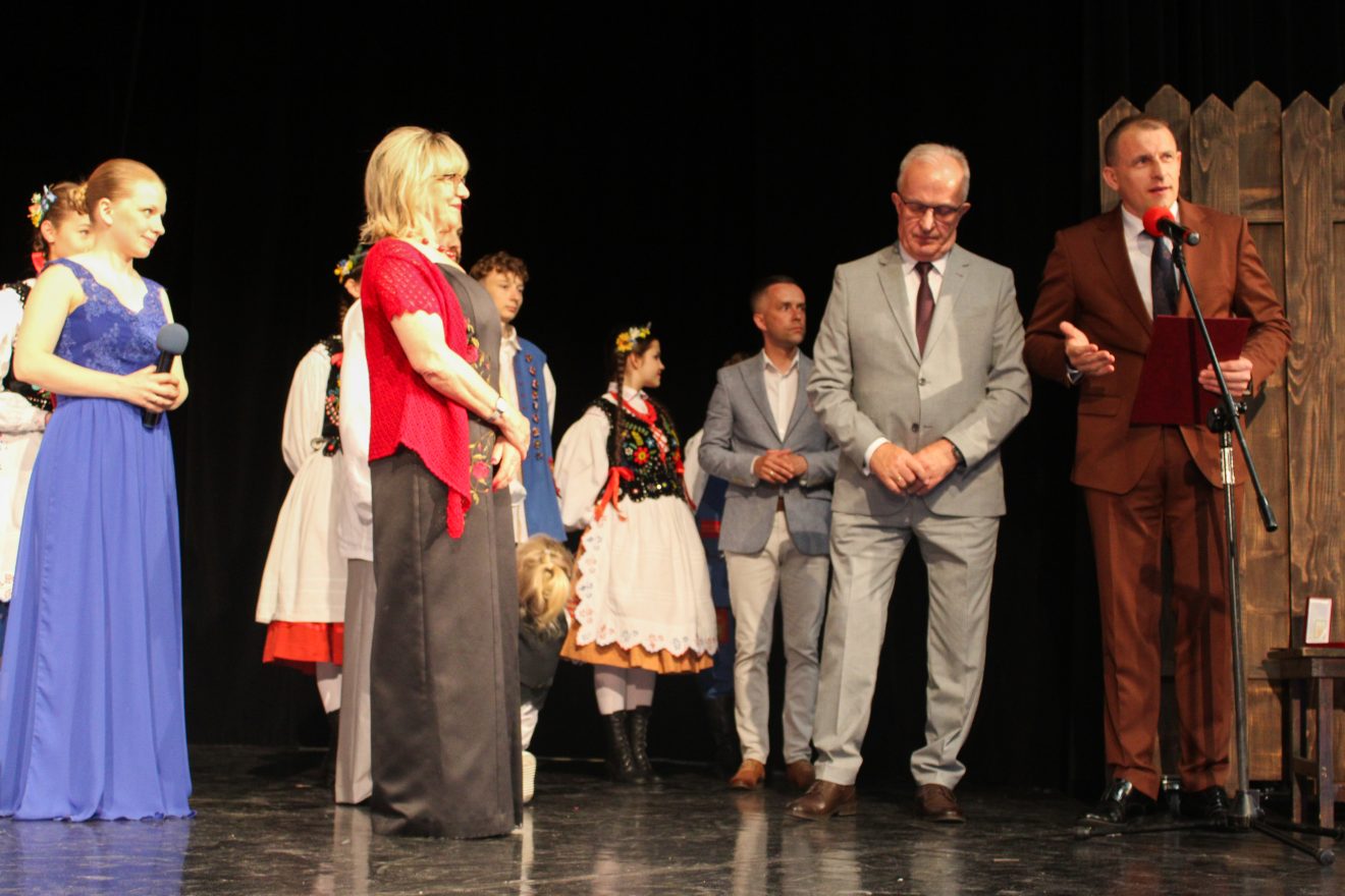 Na scenie przedstawiciele Powiatu Tomaszowskiego jeden z nich trzyma kosz upominkowy, drugi przemawia do mikrofonu. W tle dzieci w strojach ludowych z Zespołu Folklorystycznego Roztocze.