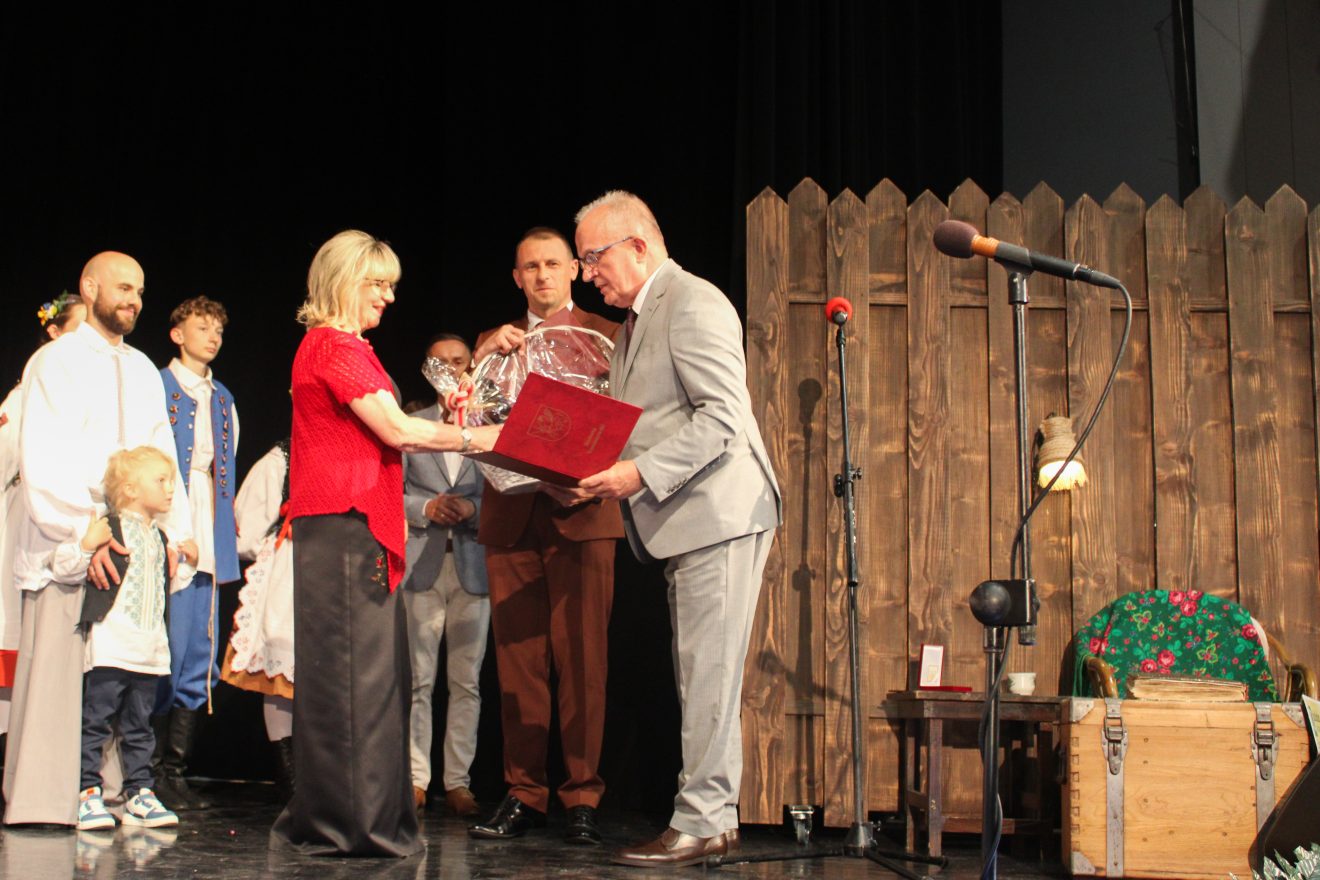 Na scenie Starosta Tomaszowski składający gratulacje na ręce założycielki i kierowniczki Zespołu Folklorystycznego Roztocze. W tle dzieci w strojach ludowych.