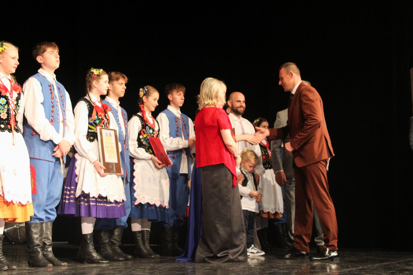 Na scenie Przewodniczący Rady Powiatu w Tomaszowie Lubelski składający gratulacje na ręce założycielki i kierowniczki Zespołu Folklorystycznego Roztocze. W tle dzieci w strojach ludowych.