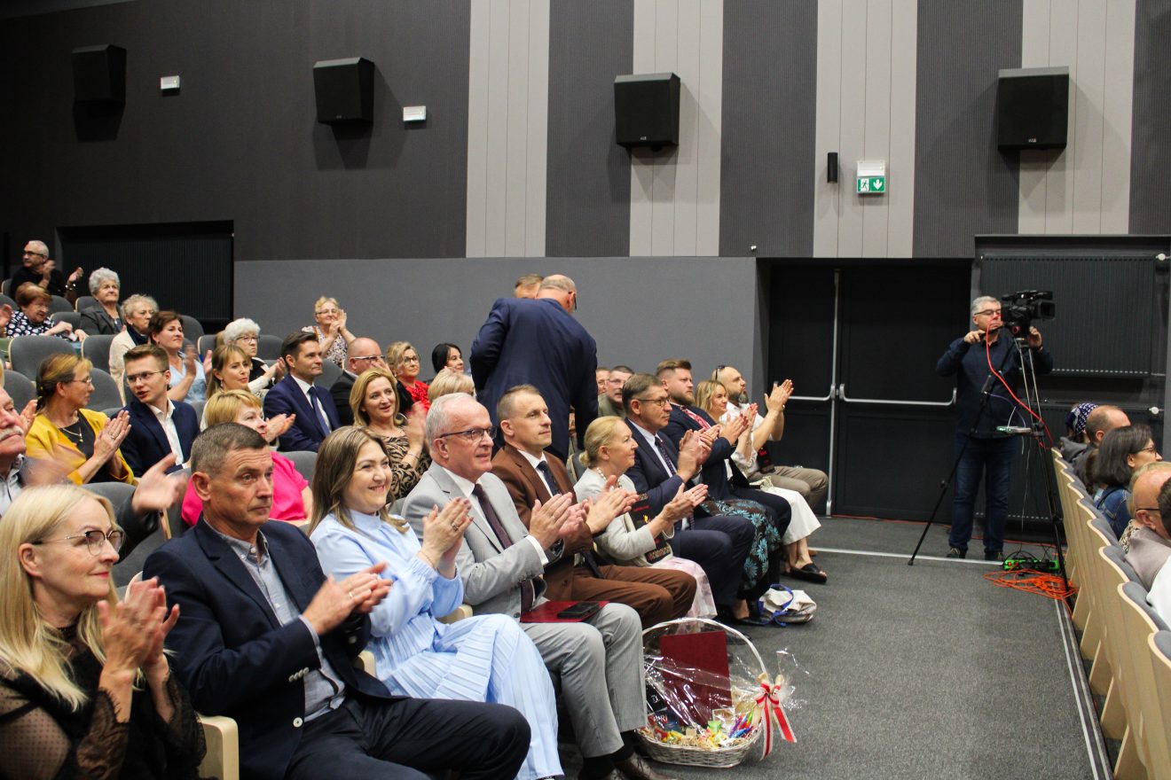 Publiczność wypełnia salę widowiskową, goście siedzący w pierwszym rzędzie biją brawo, obok nich stoi kosz upominkowy; w tle kamerzysta nagrywa wydarzenie.