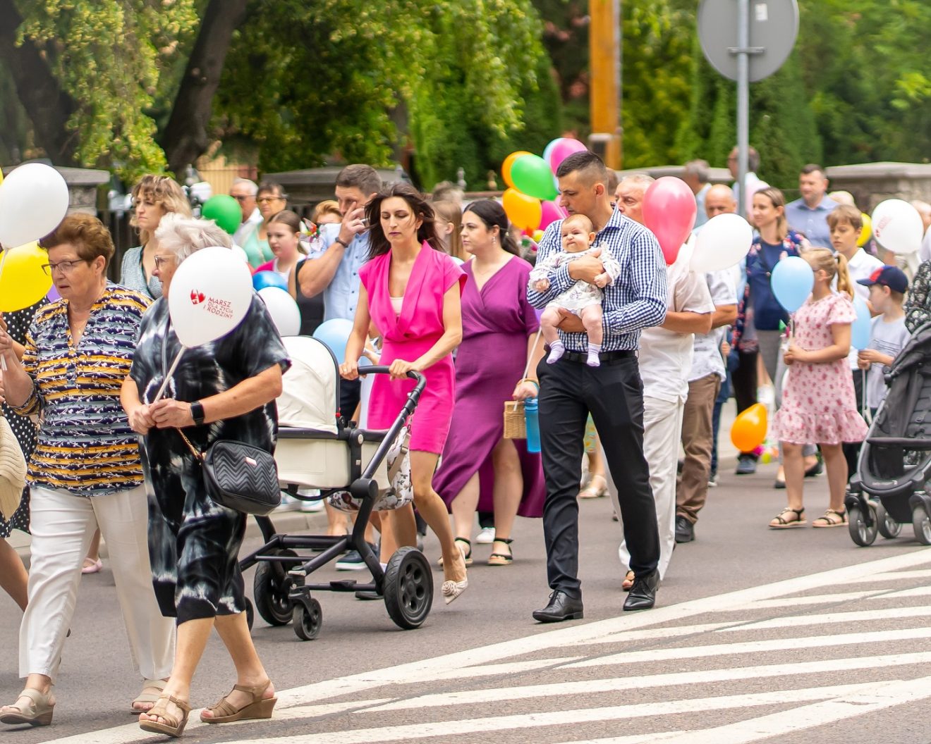 Grupa ludzi, w tym rodziny z dziećmi, maszeruje ulicą podczas wydarzenia społecznego. Wiele osób trzyma kolorowe balony, część prowadzi wózki dziecięce. Atmosfera jest radosna i rodzinna. W tle drzewa i miejska zabudowa.