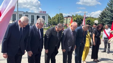 Grupa osób w oficjalnych strojach pochyla głowy w geście pamięci podczas uroczystości rocznicowych na miejskim rynku w Tomaszowie Lubelskim. W tle widoczne są flagi, budynki i niebo z chmurami.