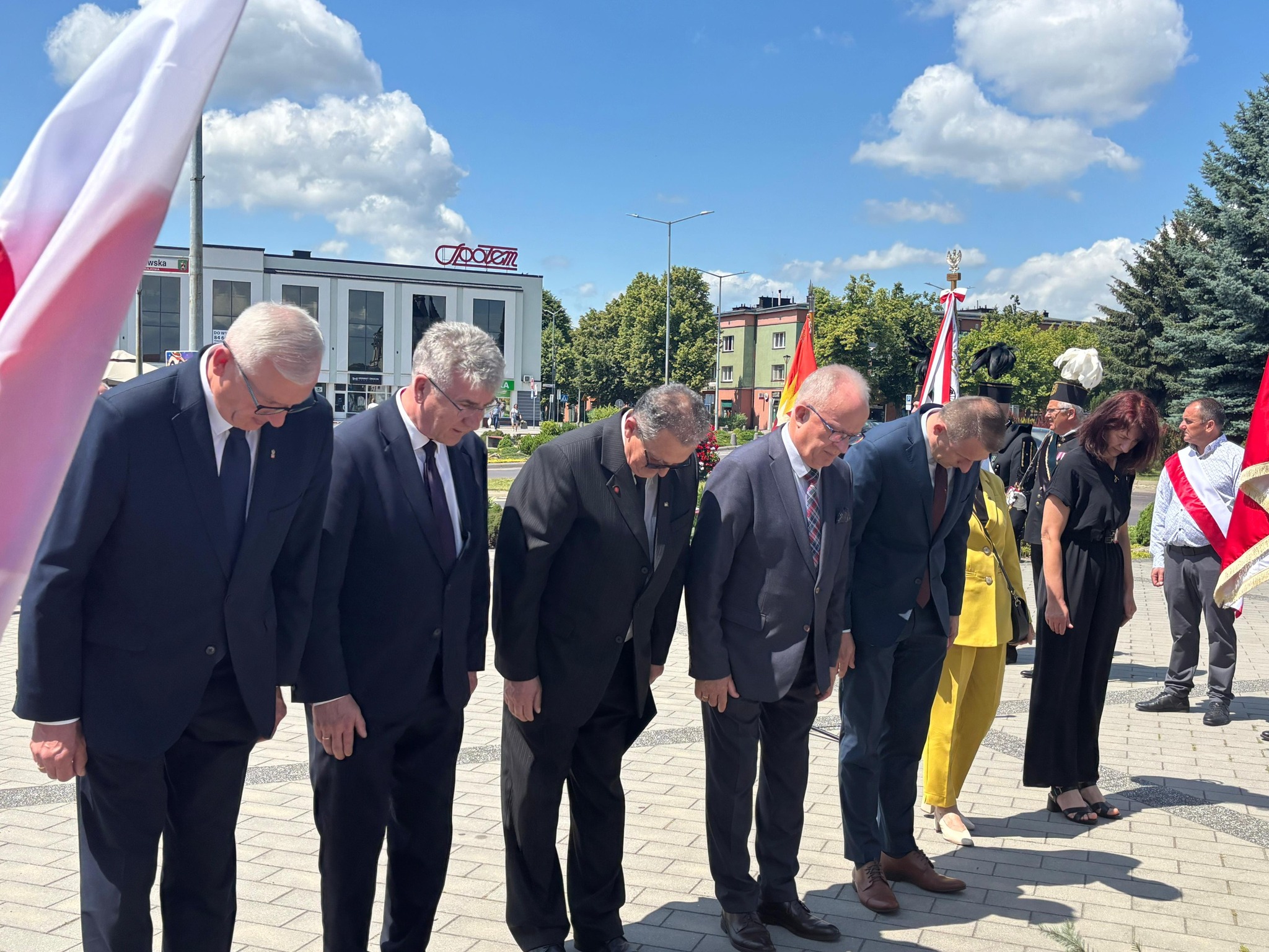 Grupa osób w oficjalnych strojach pochyla głowy w geście pamięci podczas uroczystości rocznicowych na miejskim rynku w Tomaszowie Lubelskim. W tle widoczne są flagi, budynki i niebo z chmurami.