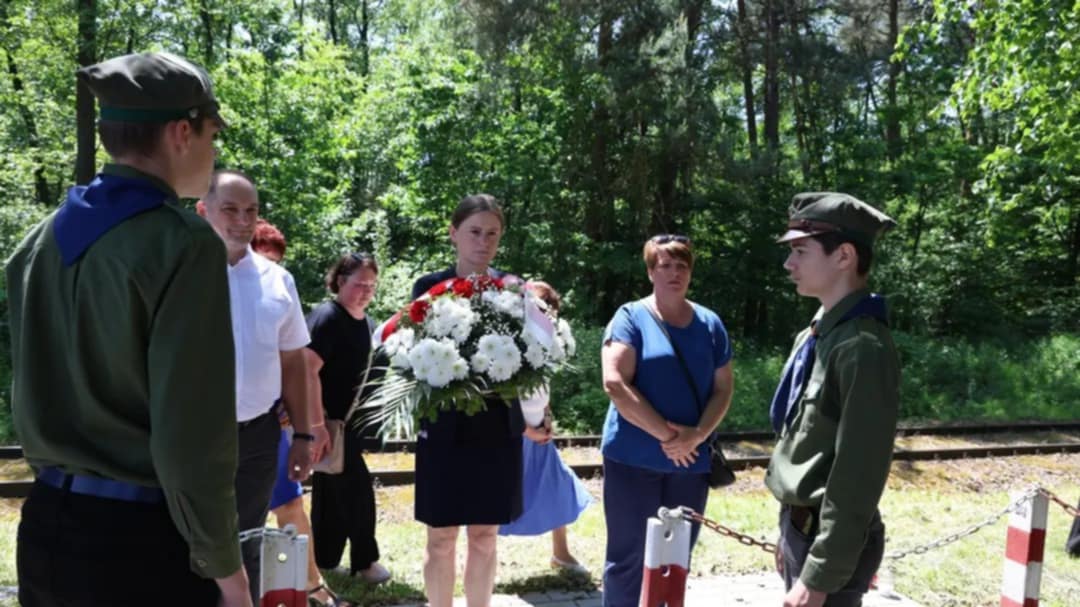 Grupa osób, w tym harcerze i przedstawicielka władz powiatowych, stoi w ciszy przed miejscem pamięci. Kobieta w centrum trzyma biało-czerwony wieniec. W tle widać tory kolejowe i otaczający teren leśny. Scena symbolizuje chwilę zadumy i hołdu dla ofiar zbrodni UPA.