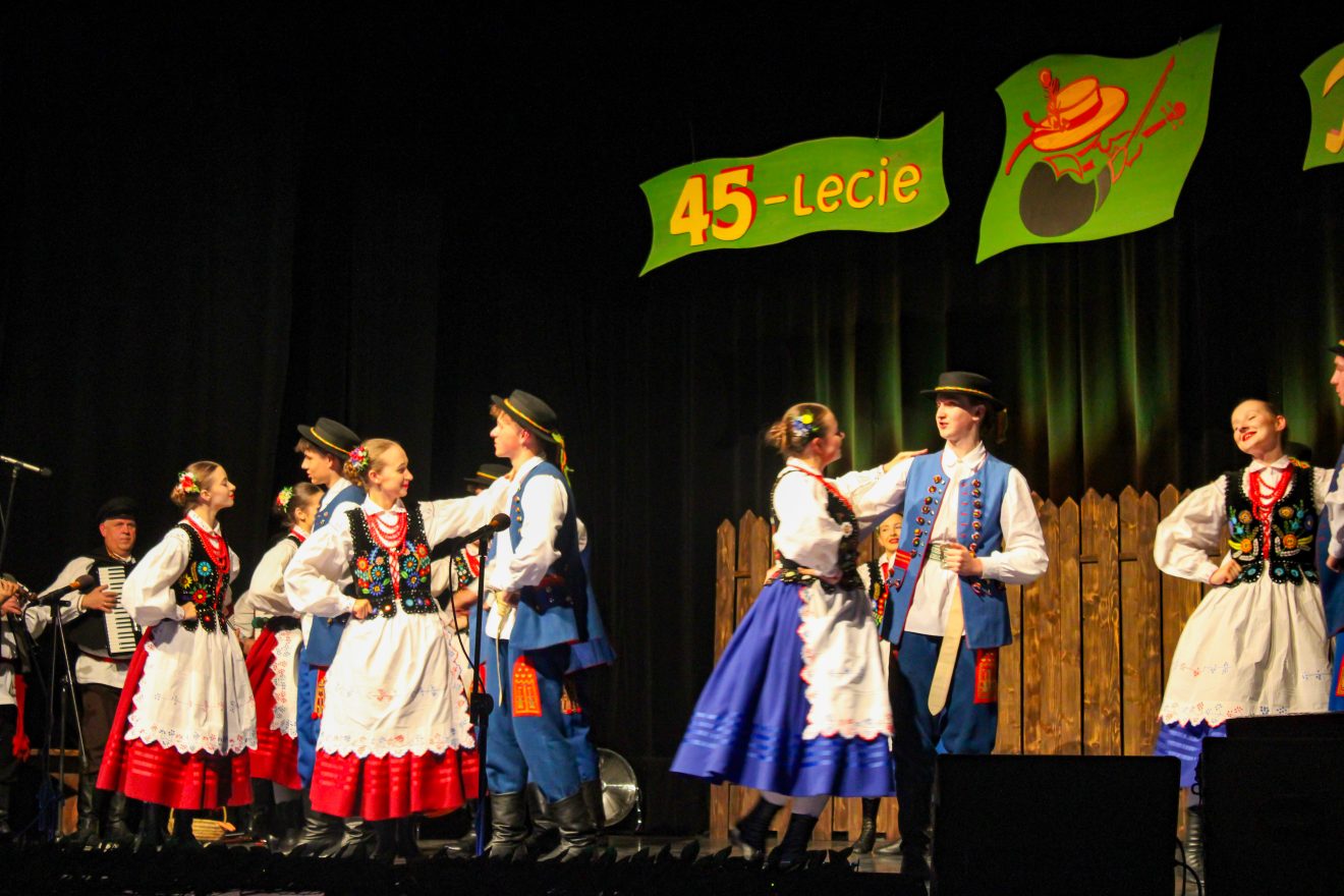 Młodzież z zespołu w strojach ludowych tańczący na scenie.