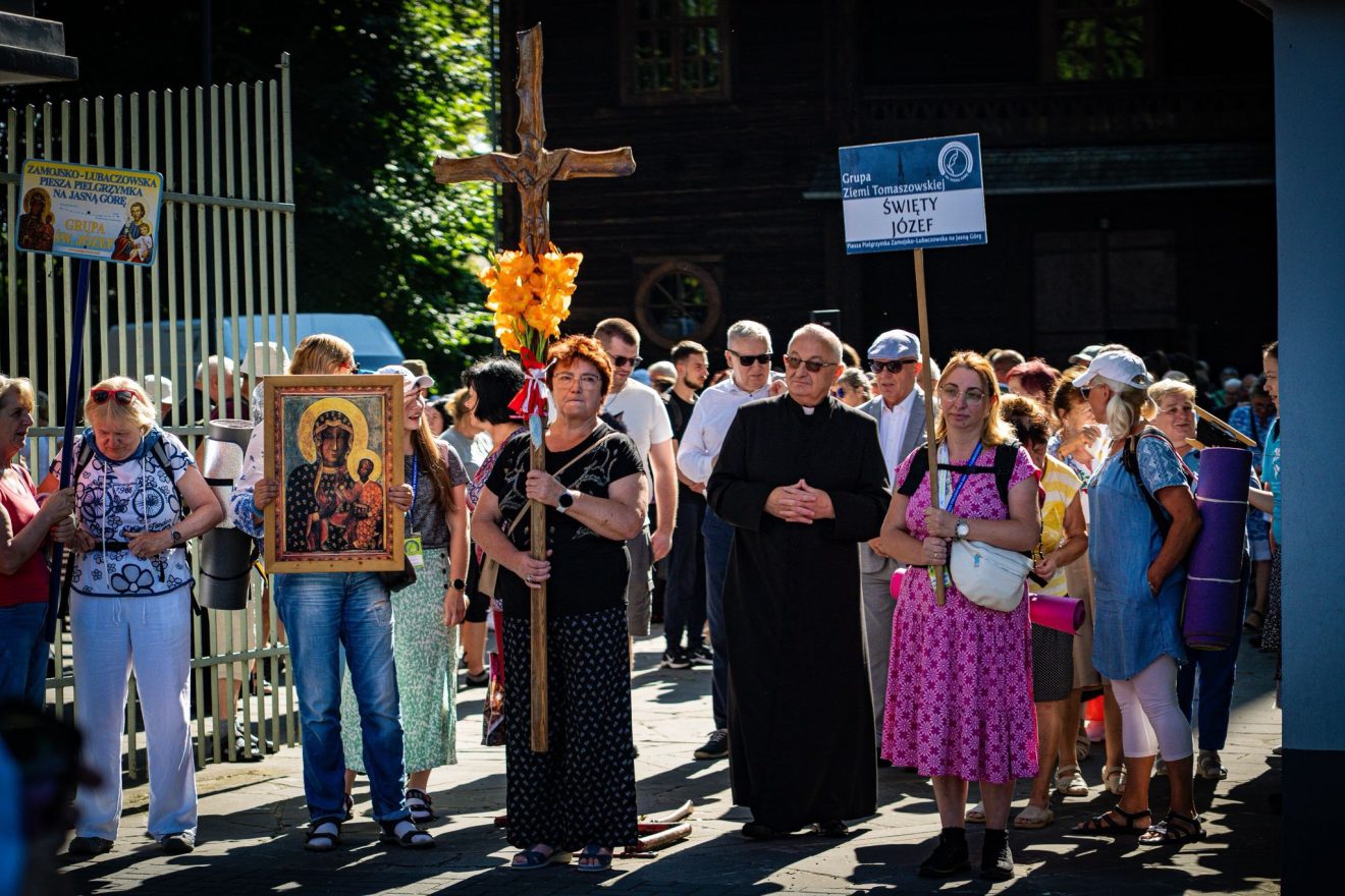Grupa pielgrzymów wyrusza pieszo z Tomaszowa Lubelskiego. Na czele niesiony jest krzyż. W tle kościół.