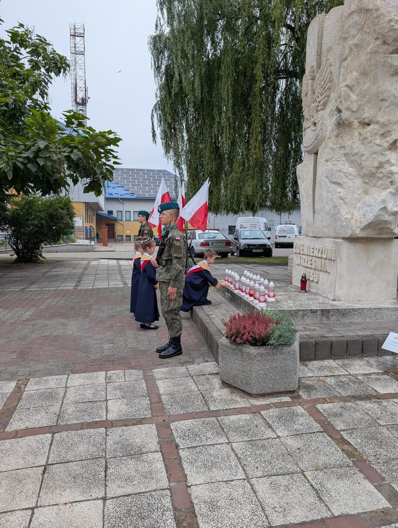 Pomnik „Żołnierzom Września 1939 r.”, przed którym stoi warta honorowa zwrócona tyłem do pomnika. U jego stóp złożone są znicze upamiętniające poległych obrońców Ojczyzny. W tle flagi biało-czerwone.