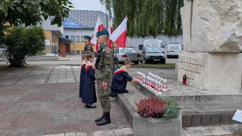Pomnik „Żołnierzom Września 1939 r.”, przed którym stoi warta honorowa zwrócona tyłem do pomnika. U jego stóp złożone są znicze upamiętniające poległych obrońców Ojczyzny. W tle flagi biało-czerwone.