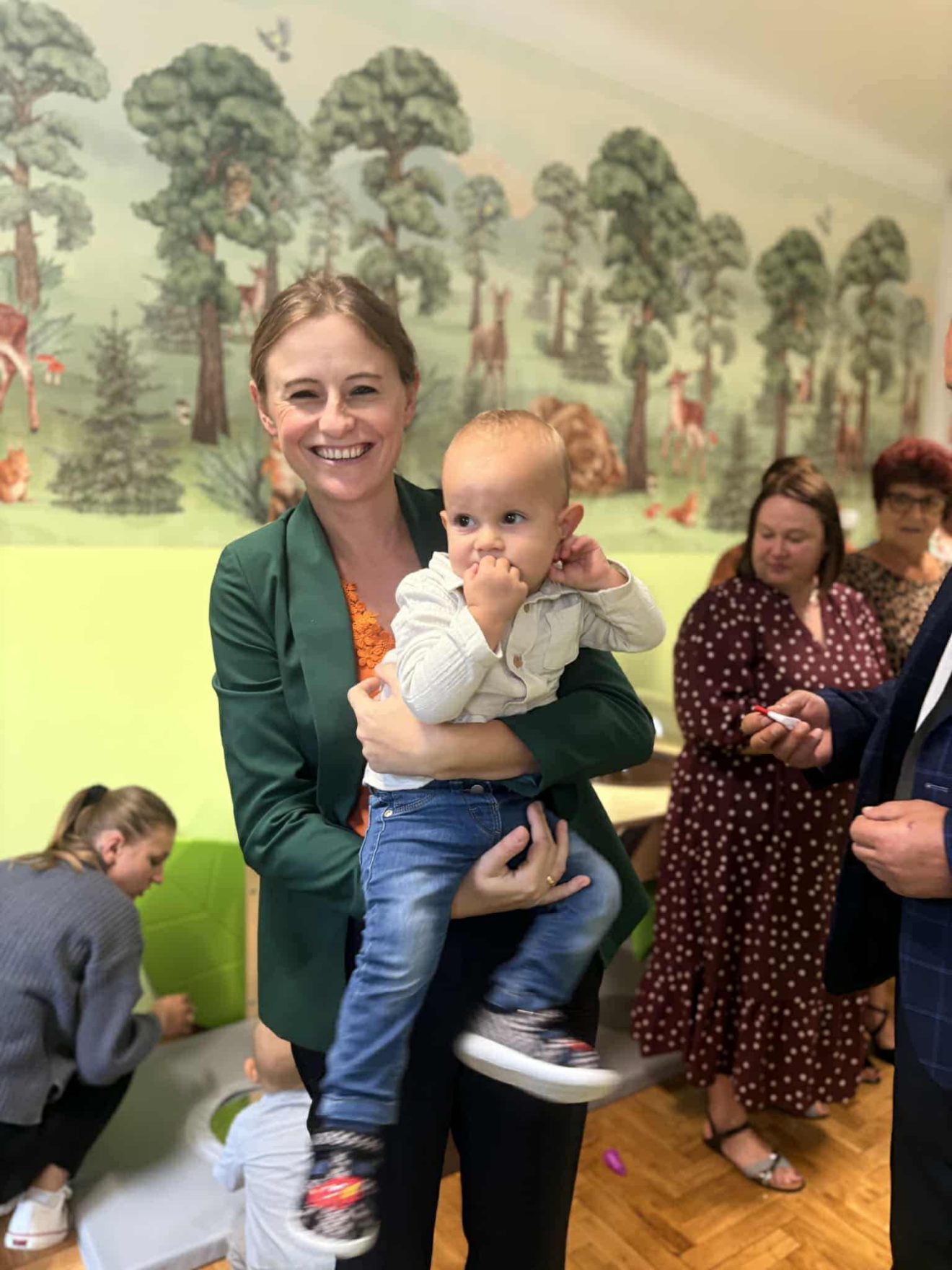 Wiceprzewodnicząca Rady Powiatu Aleksandra Wołoszyn-Banaś uśmiecha się, trzymając na rękach jedno z dzieci podczas otwarcia Punktu Dziennych Opiekunów ‘Promyczek’