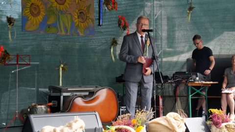 Starosta Henryk Karwan stoi na scenie przy mikrofonie i przemawia do zgromadzonych uczestników Dożynek w Pawłówce. W tle widać barwne dekoracje