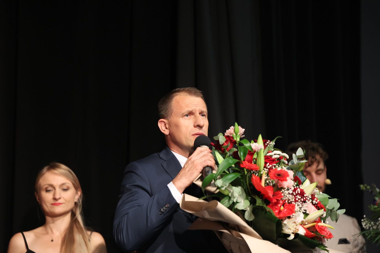Na scenie stoją członkowie Tomaszowskiego Zespołu Teatralnego, a przewodniczący Rady Powiatu Tomaszowskiego Witold Pitura, trzymając bukiet kwiatów i mikrofon, wygłasza przemówienie