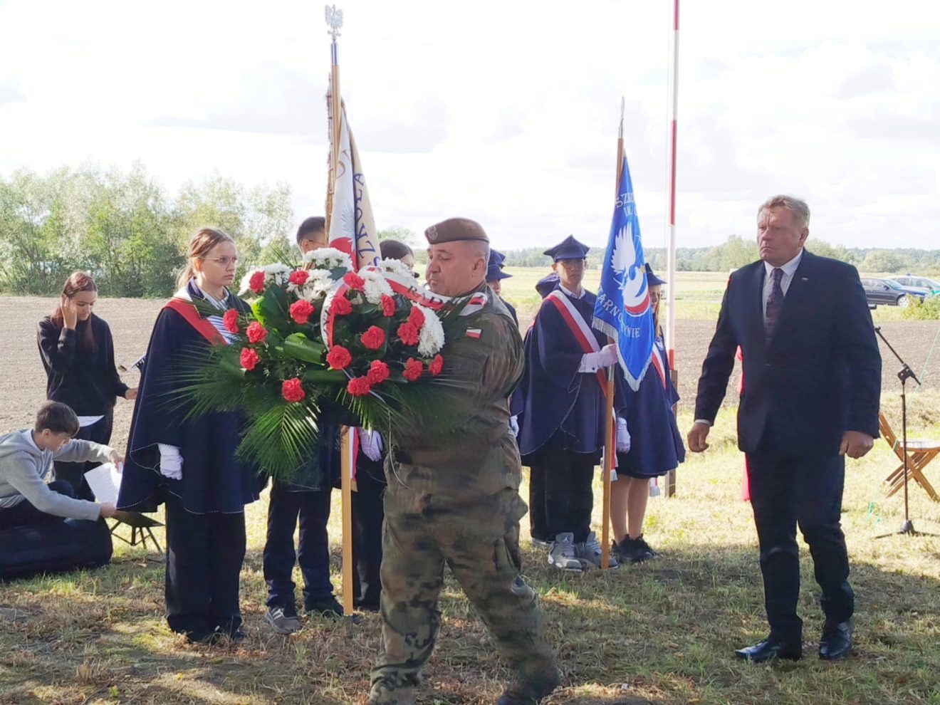 Żołnierz niesie wieniec biało-czerwonych kwiatów, aby złożyć go na grobie Bronisława Skowrońskiego, a za nim idzie Wiesław Cielica uczestniczący w uroczystości