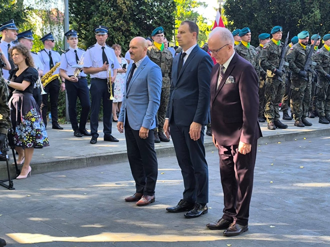Trzech przedstawicieli władz powiatu oddaje hołd po złożeniu wieńca. W tle widoczna orkiestra strażacka i żołnierze.