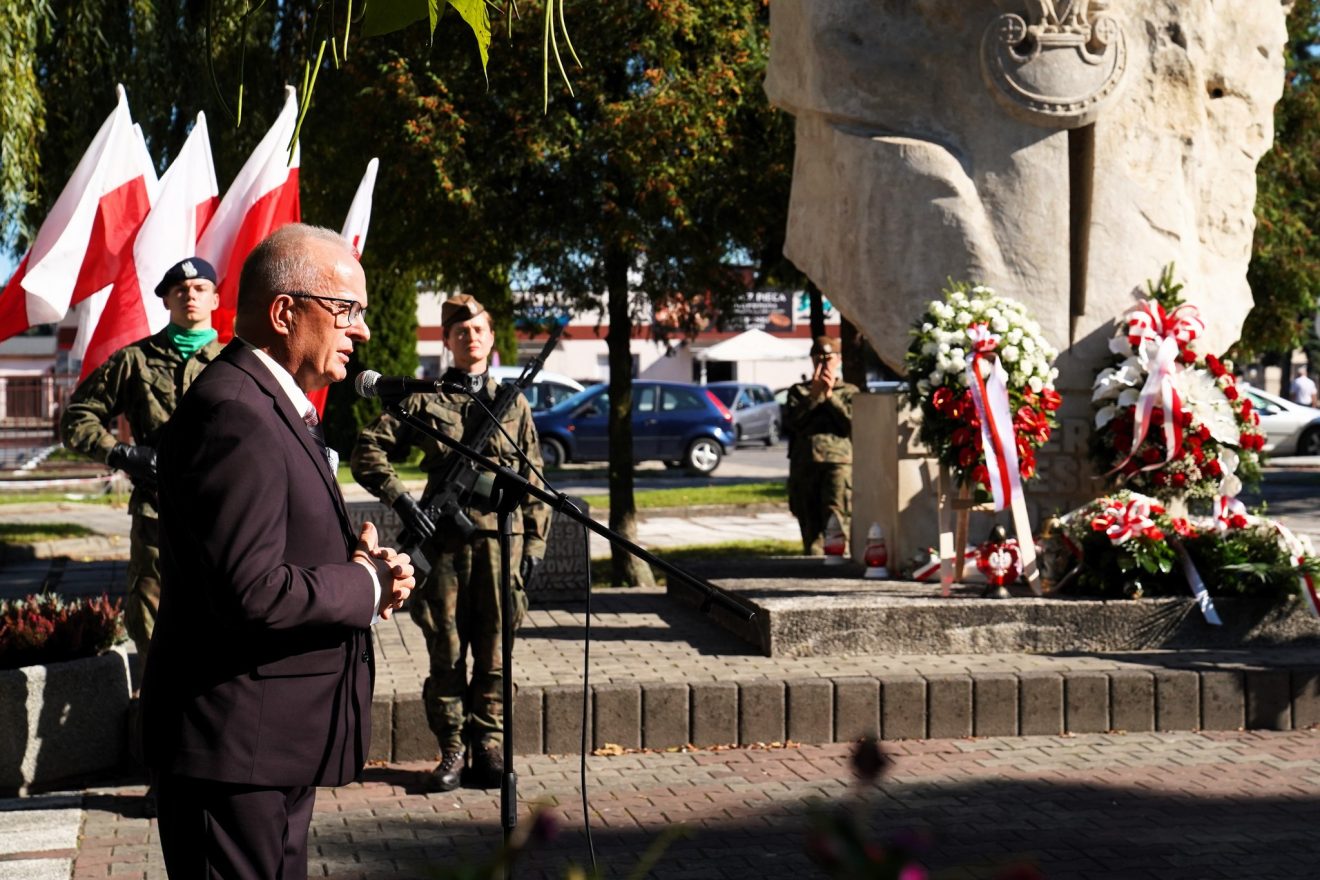Starosta podczas przemówienia przy mikrofonie; w tle widoczne flagi narodowe, pomnik oraz złożone wieńce.