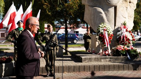 Starosta podczas przemówienia przy mikrofonie; w tle widoczne flagi narodowe, pomnik oraz złożone wieńce.