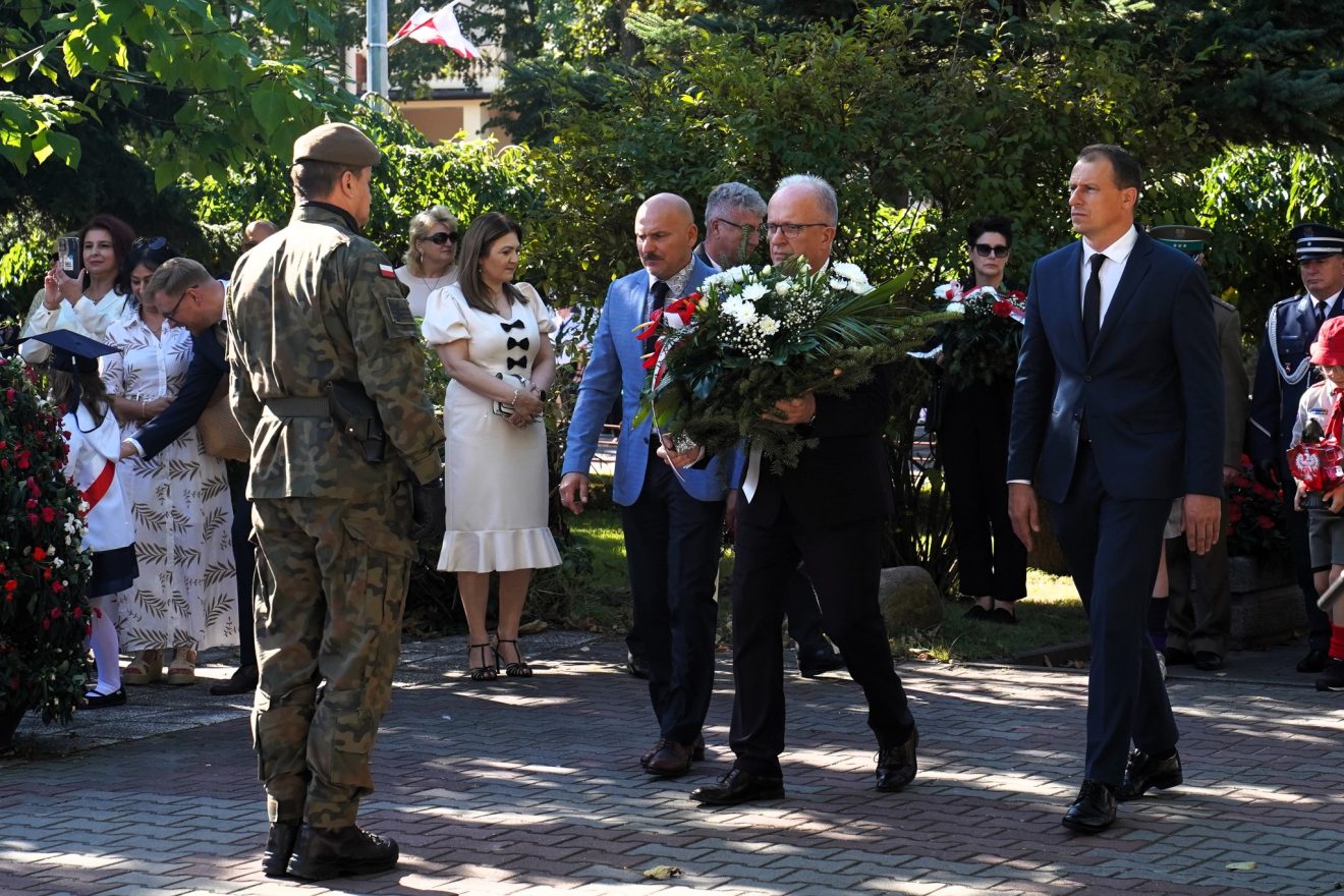 Starosta wraz z Przewodniczącym Rady Powiatu i Wicestarostą uczestniczą w ceremonii złożenia biało-czerwonego wieńca. W tle inni uczestnicy uroczystości.