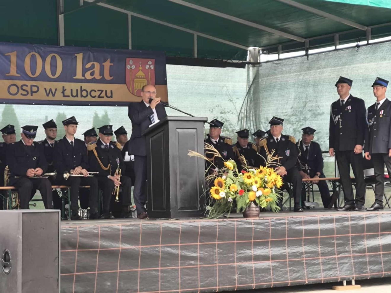 Dariusz Czop, członek Zarządu Powiatu, przemawia na jubileuszu 100-lecia OSP Łubcze, w tle siedzą strażacy.