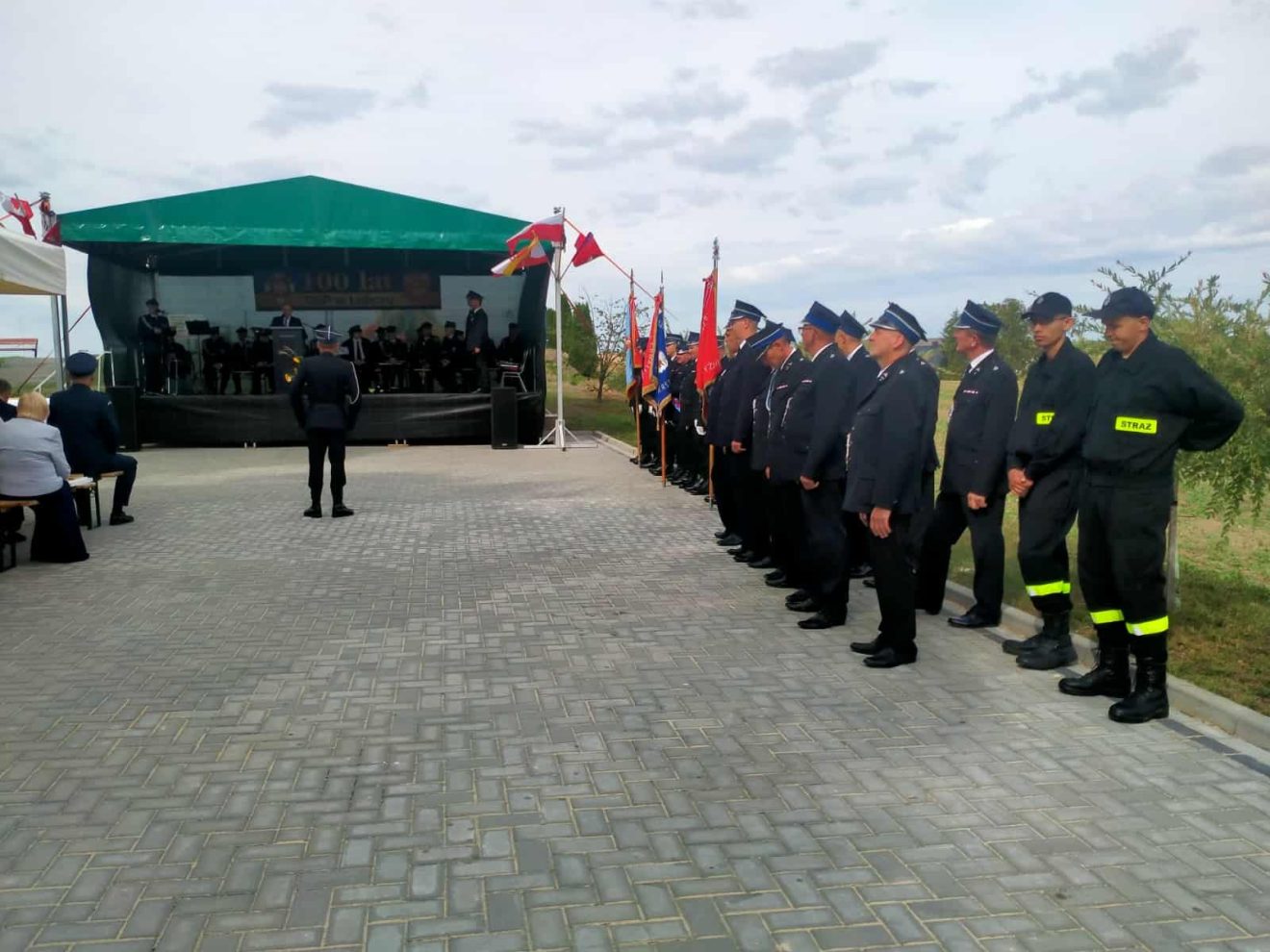 Dariusz Czop, członek Zarządu Powiatu, przemawia na scenie podczas jubileuszu 100-lecia OSP Łubcze, na scenie siedzą i przed sceną po prawej stronie stoją strażacy.