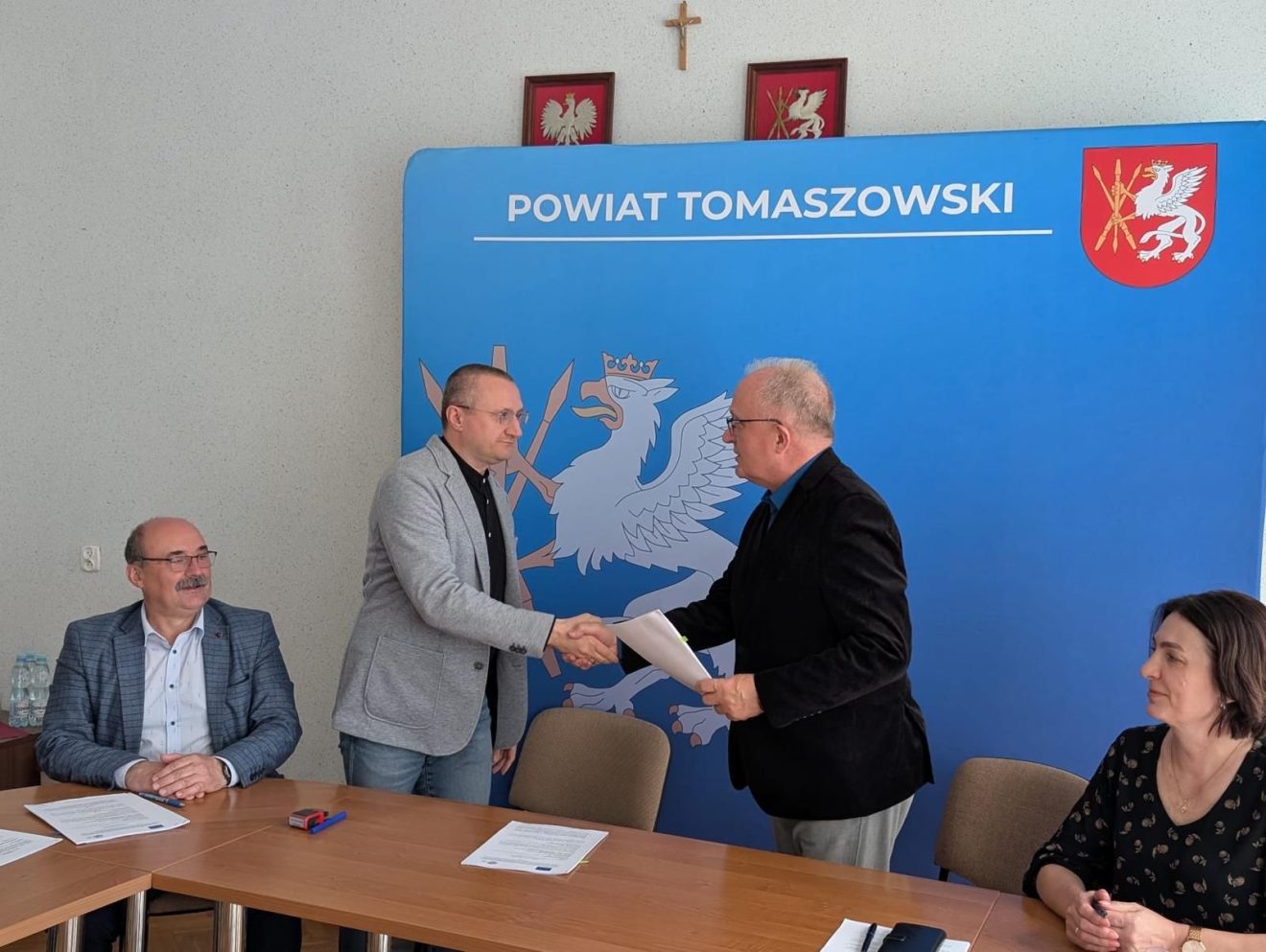 Na zdjęciu widać moment podpisania umowy – Starosta Tomaszowski podaje rękę przedstawicielowi wykonawcy na tle ścianki z napisem „Powiat Tomaszowski”. Przy stole siedzą również pozostali uczestnicy spotkania.