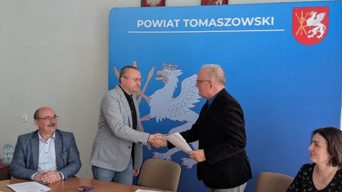 Na zdjęciu widać moment podpisania umowy – Starosta Tomaszowski podaje rękę przedstawicielowi wykonawcy na tle ścianki z napisem „Powiat Tomaszowski”. Przy stole siedzą również pozostali uczestnicy spotkania.