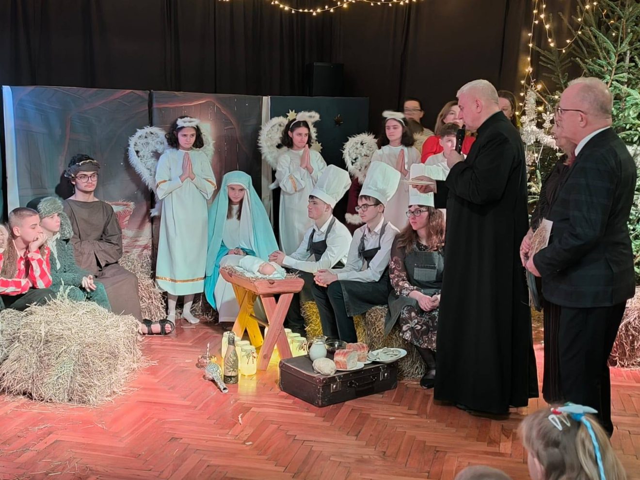 Uczniowie i zaproszeni goście podczas szkolnego Wieczoru Wigilijnego – scena jasełek z Maryją, Józefem, Dzieciątkiem Jezus i aniołami w świątecznej scenerii.
