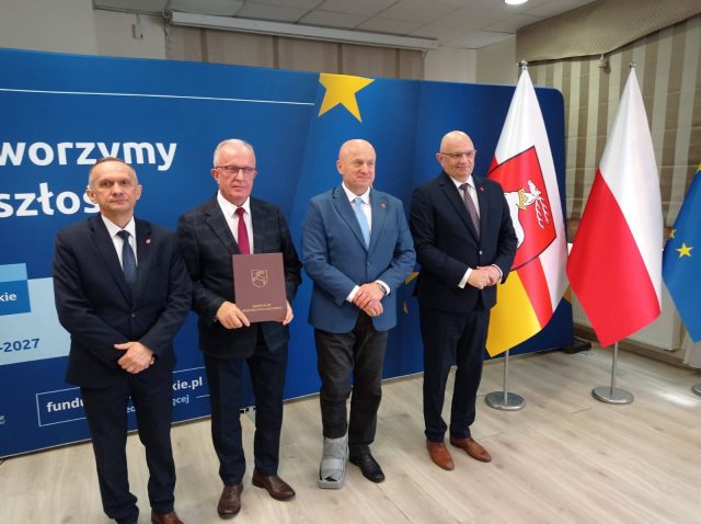 Na zdjęciu znajdują się: Henryk Karwan- Starosta Tomaszowski, Wiesław Bondyra - Dyrektor Powiatowego Urzędu Pracy, Jarosław Stawiarski - Marszałek Województwa Lubelskiego, na tle niebieskiej ścianki informacyjnej , a po prawej stronie zdjęcia znajdują się flagi: polska i urzędu marszałkowskiego.
