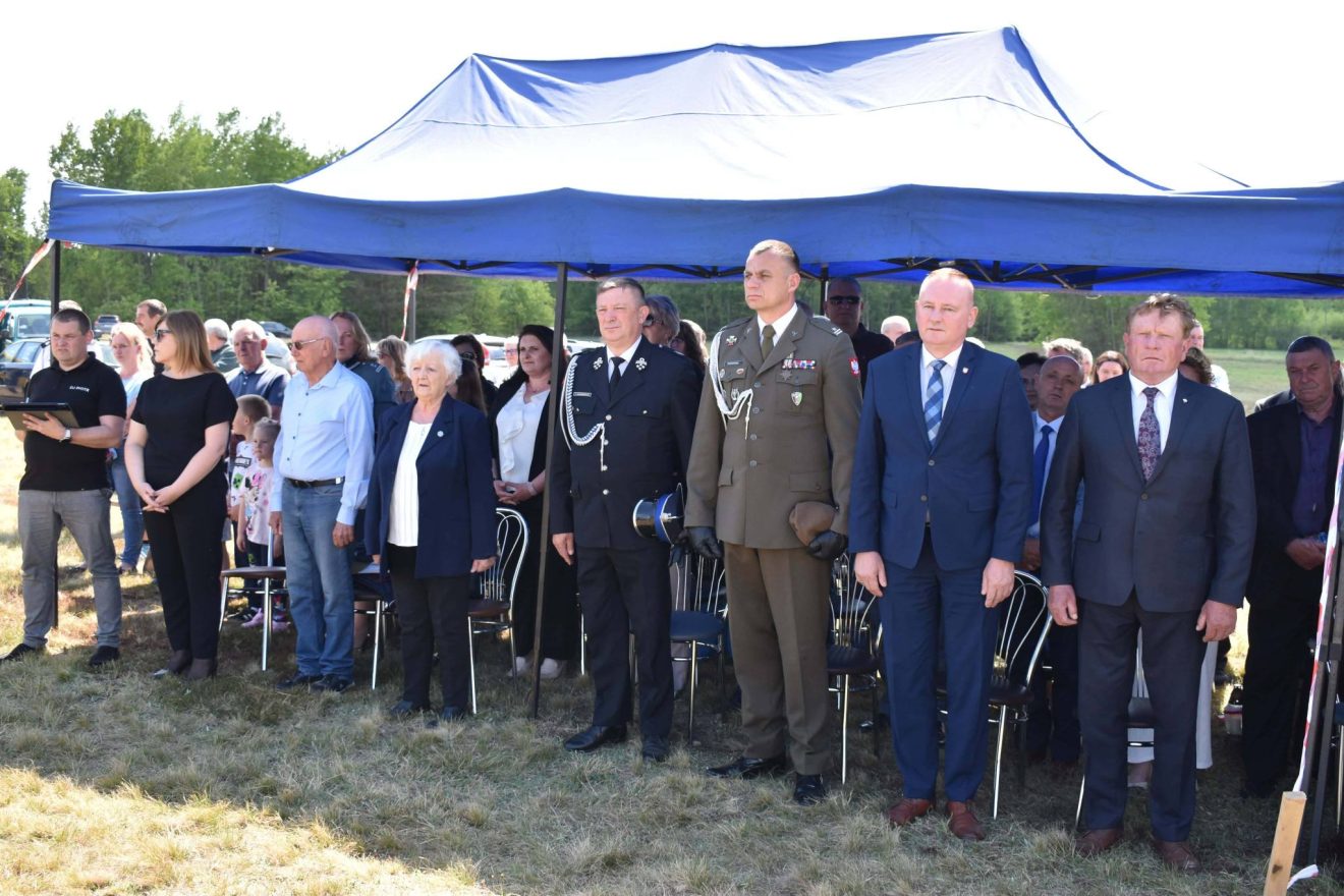 Uczestnicy uroczystości patriotycznych stoją podczas oficjalnej części obchodów pod namiotem plenerowym.
