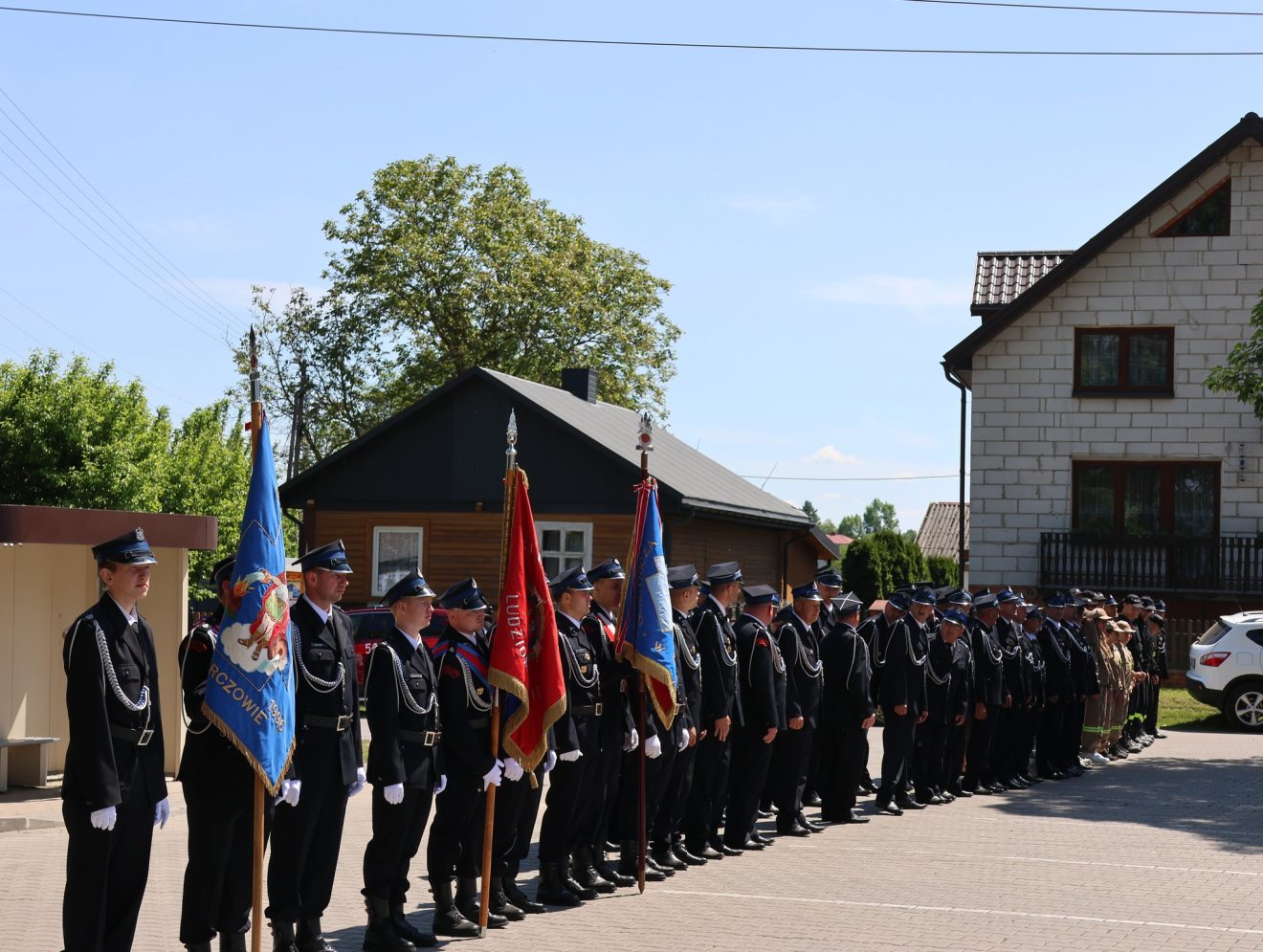 Poczty sztandarowe i strażacy stoją podczas uroczystości jubileuszowych przed remizą OSP.