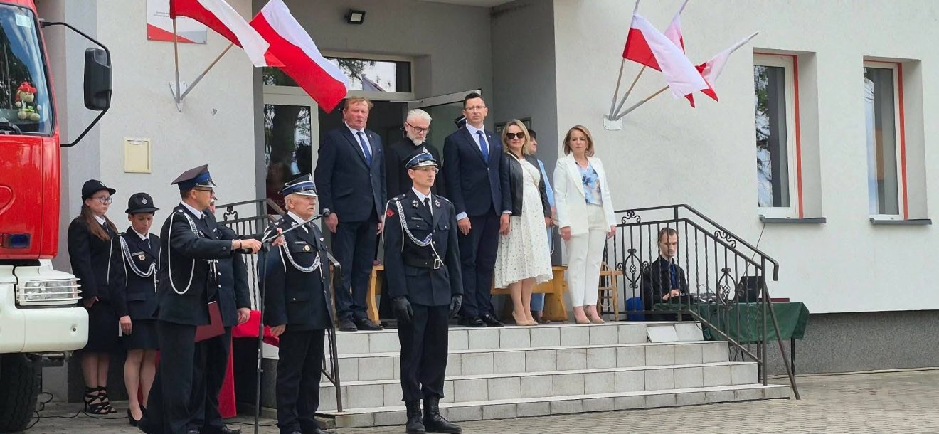 Przed budynkiem OSP podczas uroczystości jubileuszowych stoją strażacy oraz zaproszeni goście, a nad wejściem powiewają biało-czerwone flagi.