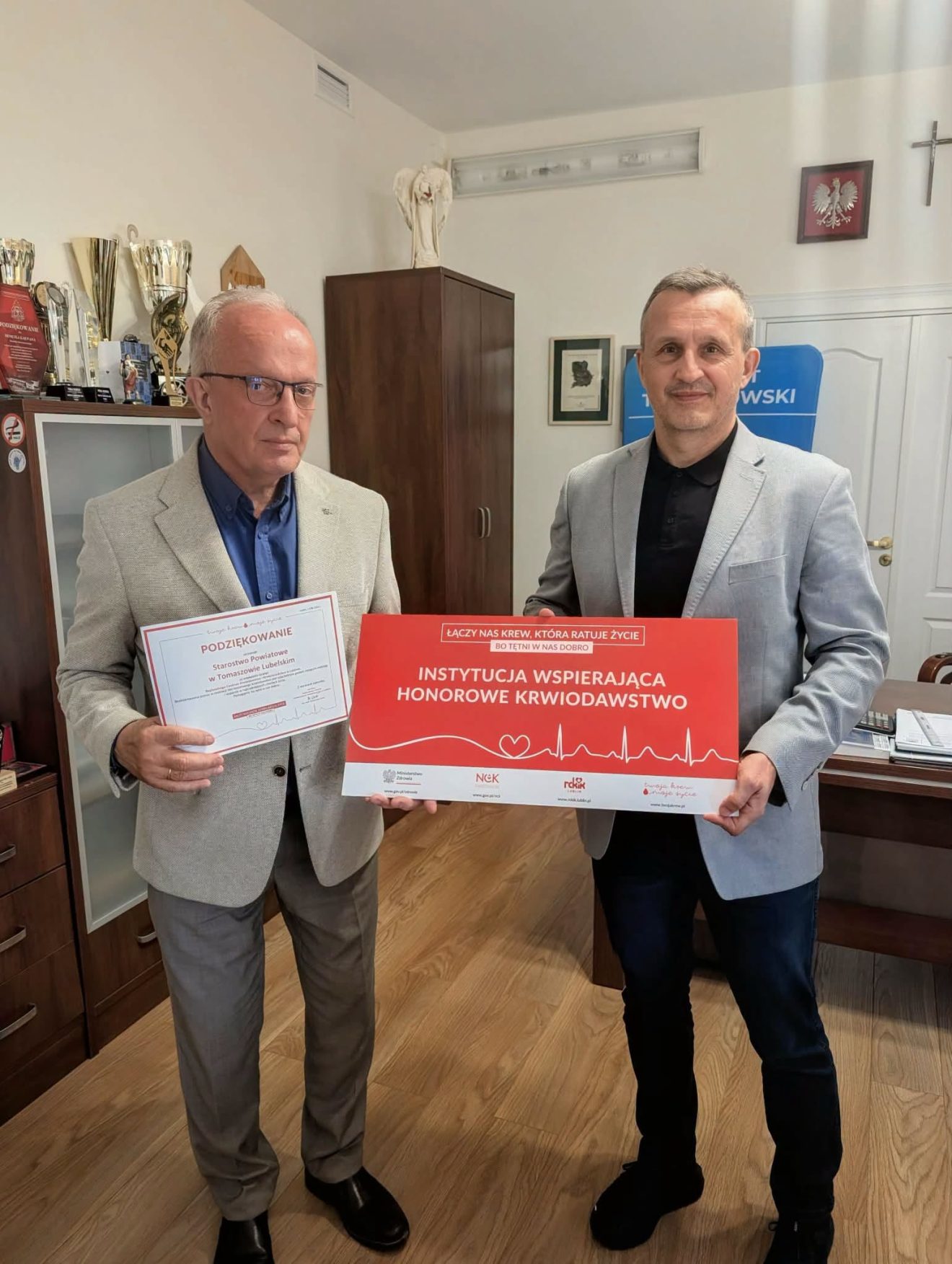 Na zdjęciu znajduje się Starosta Tomaszowski, który trzyma podziękowania za przeprowadzane akcje krwiodawstwa w Starostwie oraz przedstawiciel Regionalnego Centrum Krwiodawstwa i Krwiolecznictwa który trzyma w rękach tablicę z informacją o tom ,że Starostwo Powiatowe w Tomaszowie Lubelskim jest Instytucją wspierającą krwiodawstwo.