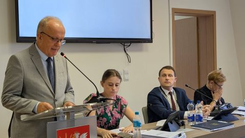 Starosta Powiatu Tomaszowskiego przemawia przy mównicy podczas sesji Rady Powiatu. Przy stole prezydialnym siedzą przewodniczący obrad oraz wiceprzewodniczące.