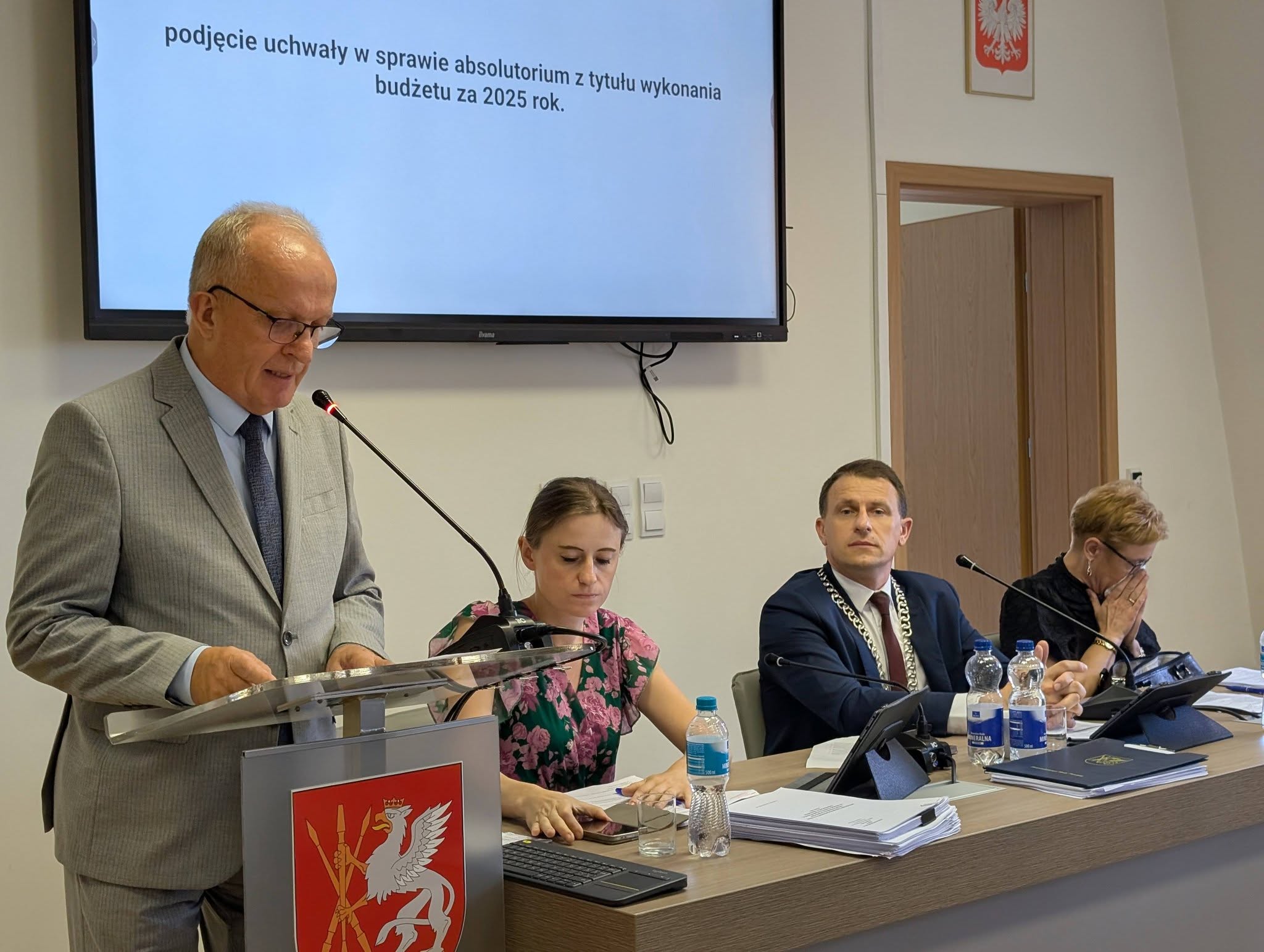 Starosta Powiatu Tomaszowskiego przemawia przy mównicy podczas sesji Rady Powiatu. Przy stole prezydialnym siedzą przewodniczący obrad oraz wiceprzewodniczące.