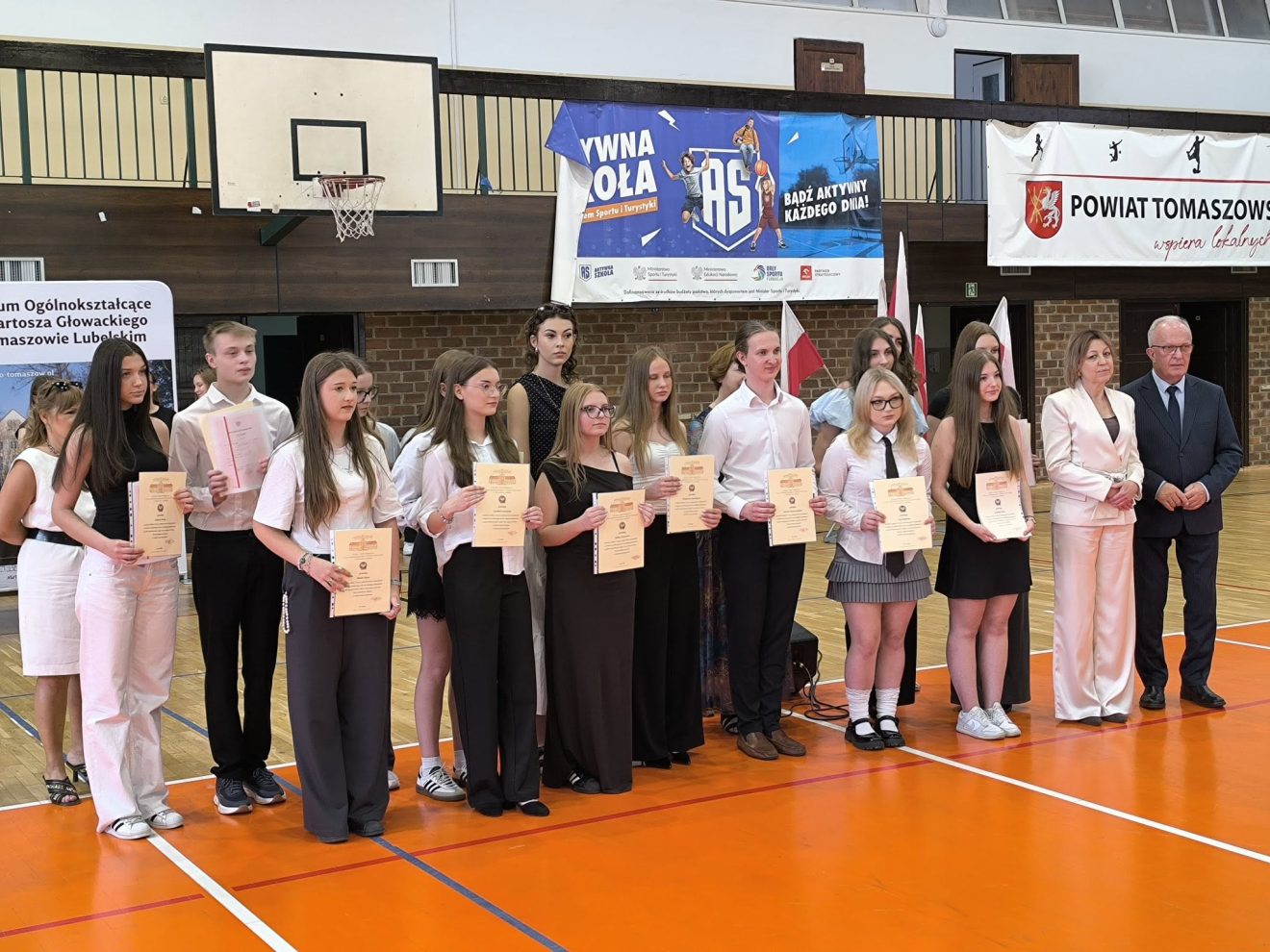 Grupa wyróżnionych uczniów pozuje z dyplomami podczas uroczystego zakończenia roku szkolnego. Obok uczniów stoją przedstawiciele szkoły oraz Starosta Tomaszowski.