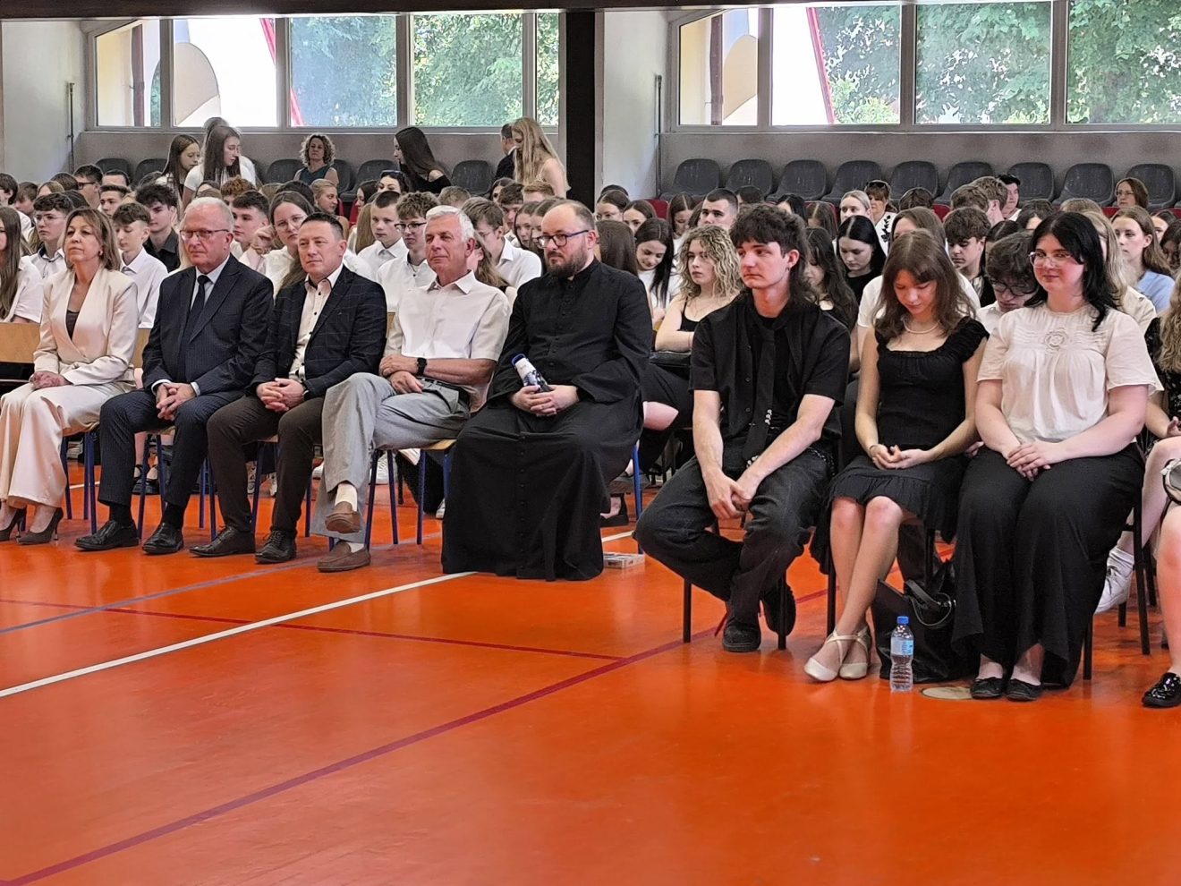 Goście, nauczyciele, rodzice i uczniowie uczestniczą w uroczystości zakończenia roku szkolnego na sali gimnastycznej.