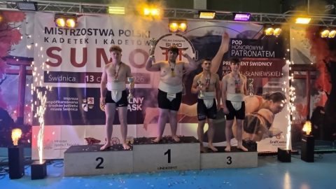 Młodzi zawodnicy sumo stoją na podium z medalami i pucharami podczas dekoracji zwycięzców Mistrzostw Polski Kadetek i Kadetów w Świdnicy. W tle widoczny jest baner zawodów oraz efekty świetlne i pirotechniczne.