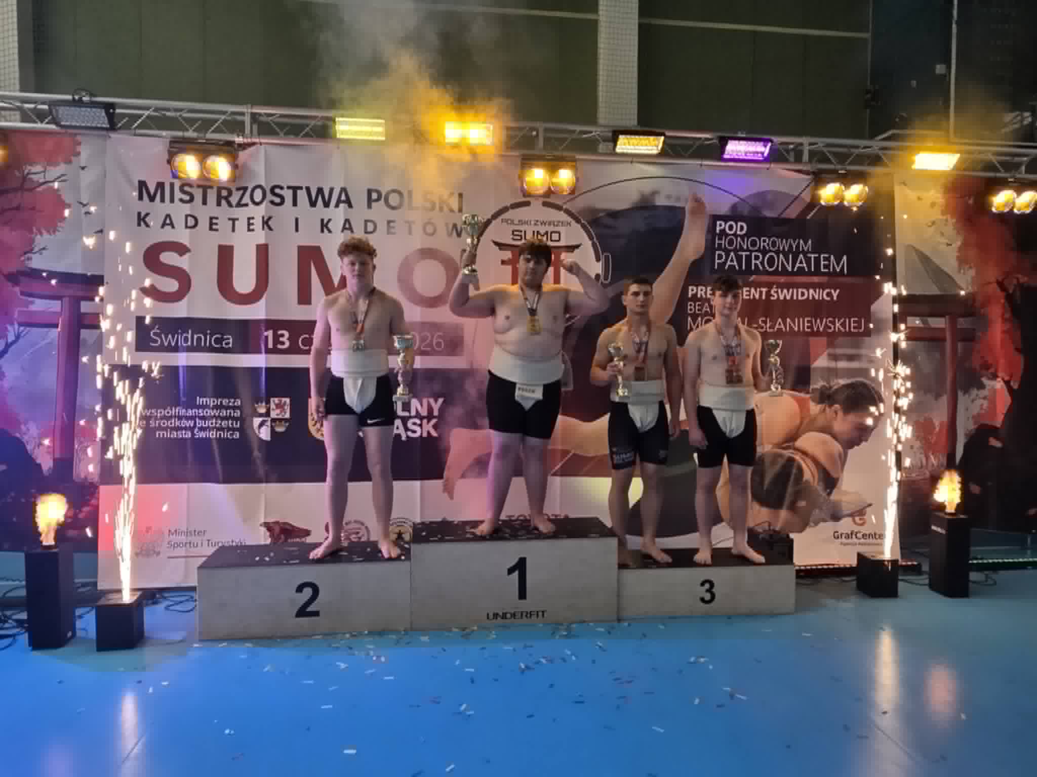 Młodzi zawodnicy sumo stoją na podium z medalami i pucharami podczas dekoracji zwycięzców Mistrzostw Polski Kadetek i Kadetów w Świdnicy. W tle widoczny jest baner zawodów oraz efekty świetlne i pirotechniczne.