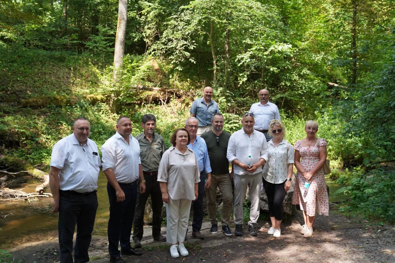 Uczestnicy spotkania pozują do wspólnego zdjęcia nad leśnym potokiem w otoczeniu gęstego lasu.