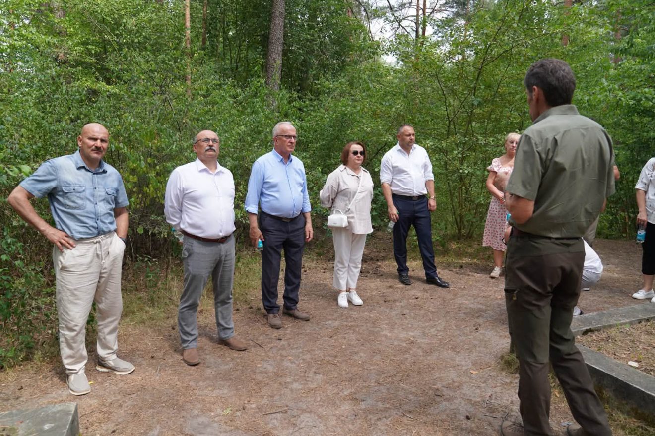 Grupa przedstawicieli samorządów i leśników stoi na leśnej ścieżce podczas wizyty terenowej i wysłuchuje informacji przewodnika.