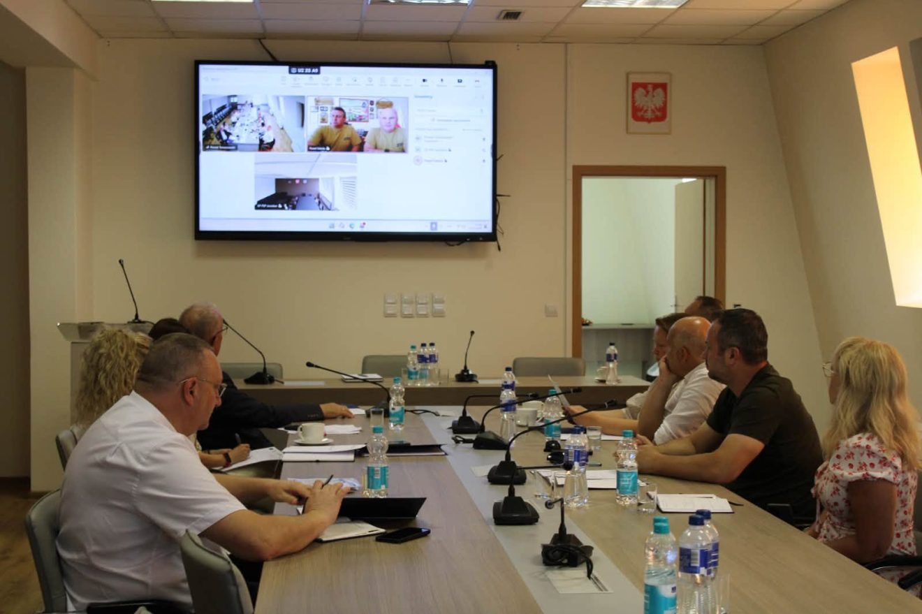 Przedstawiciele samorządów uczestniczą w roboczym spotkaniu przy stole konferencyjnym, a na ekranie widoczni są uczestnicy łączący się zdalnie.