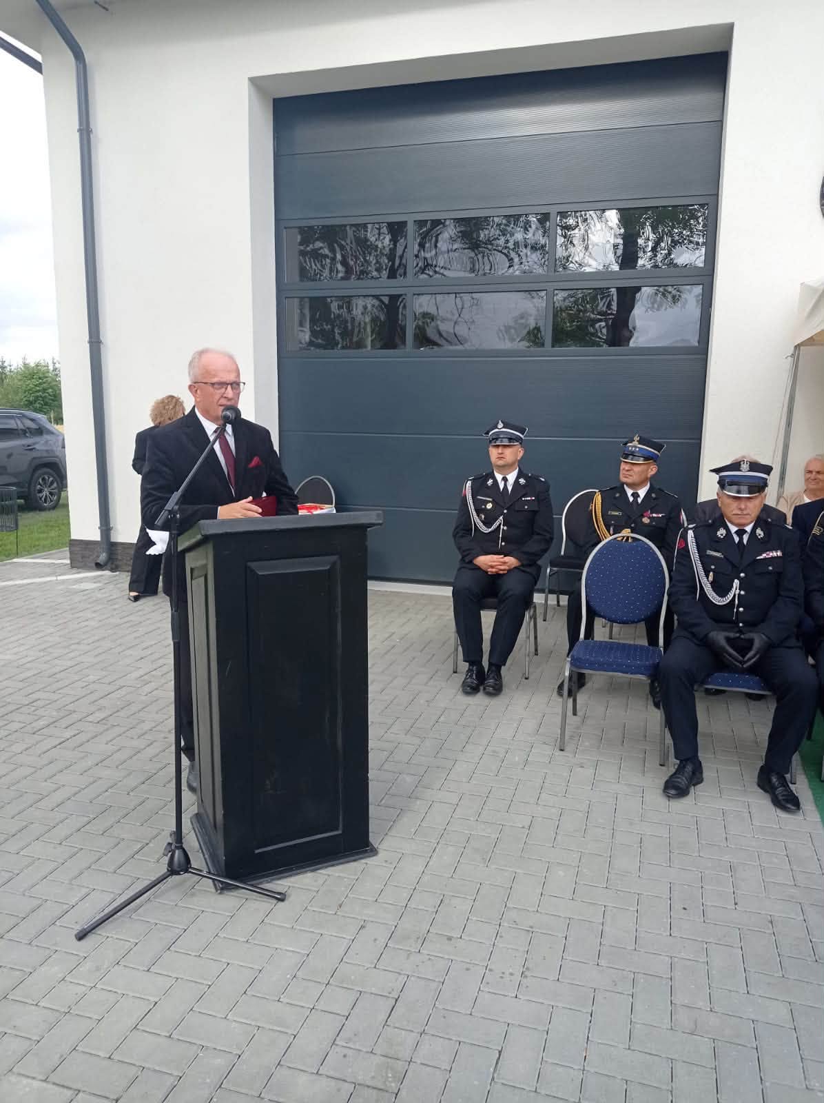 Starosta Tomaszowski wygłasza przemówienie przed nową remizą. Za nim siedzą strażacy w galowych mundurach.