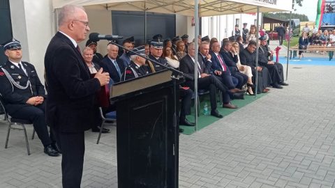 Starosta Tomaszowski przemawia przy mównicy podczas uroczystości otwarcia remizy. W tle siedzą zaproszeni goście i strażacy.