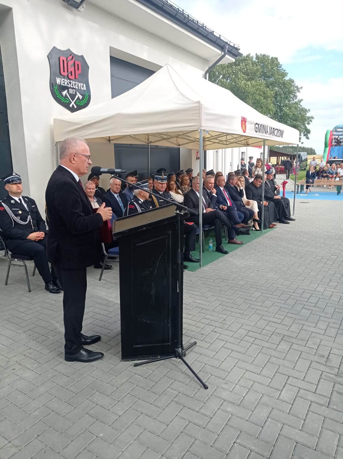Starosta Tomaszowski przemawia przy mównicy podczas uroczystości otwarcia remizy. W tle siedzą zaproszeni goście i strażacy.