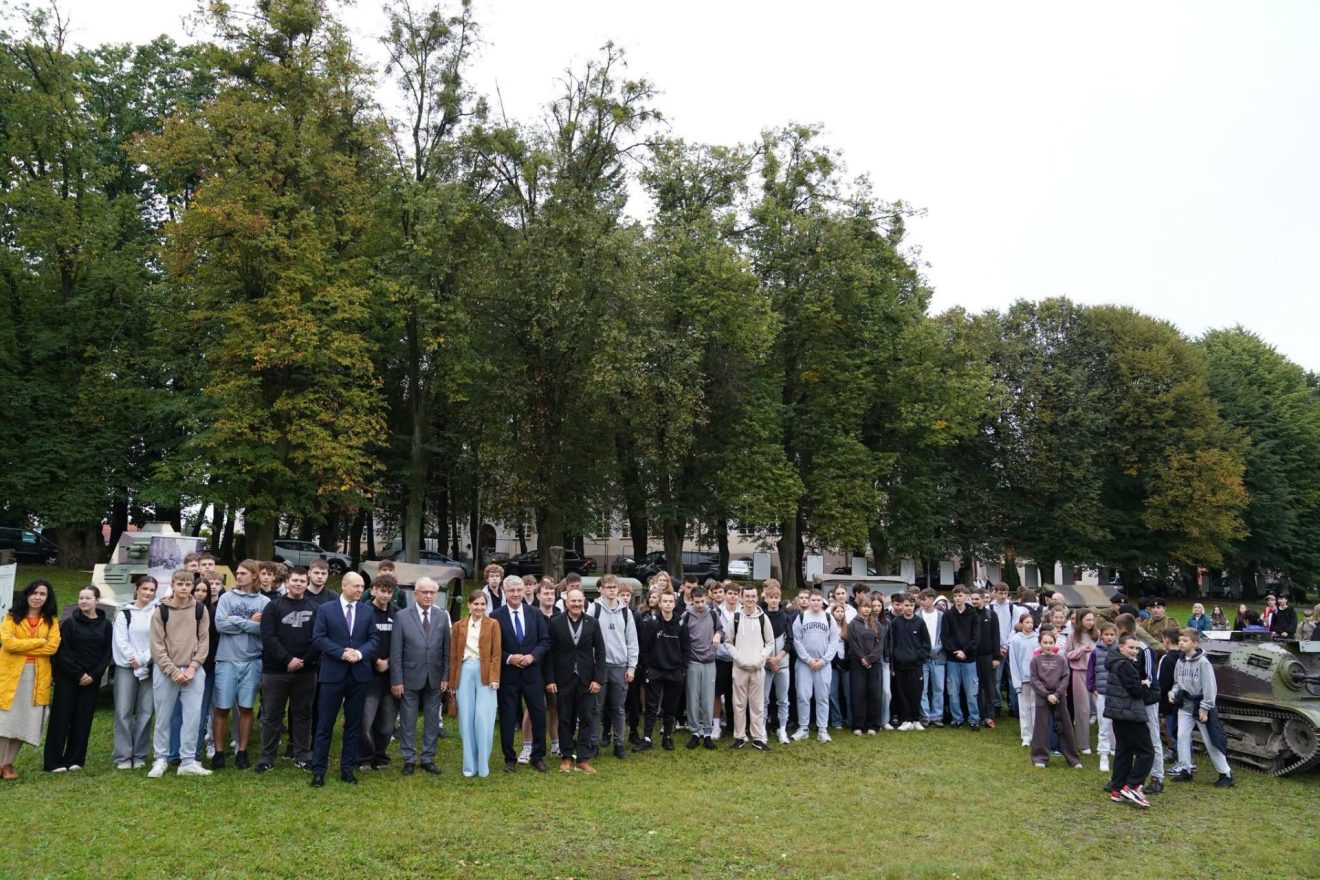 Grupowe zdjęcie młodzieży i uczestników wydarzenia „Wojenne Drogi” na tle historycznych pojazdów wojskowych.