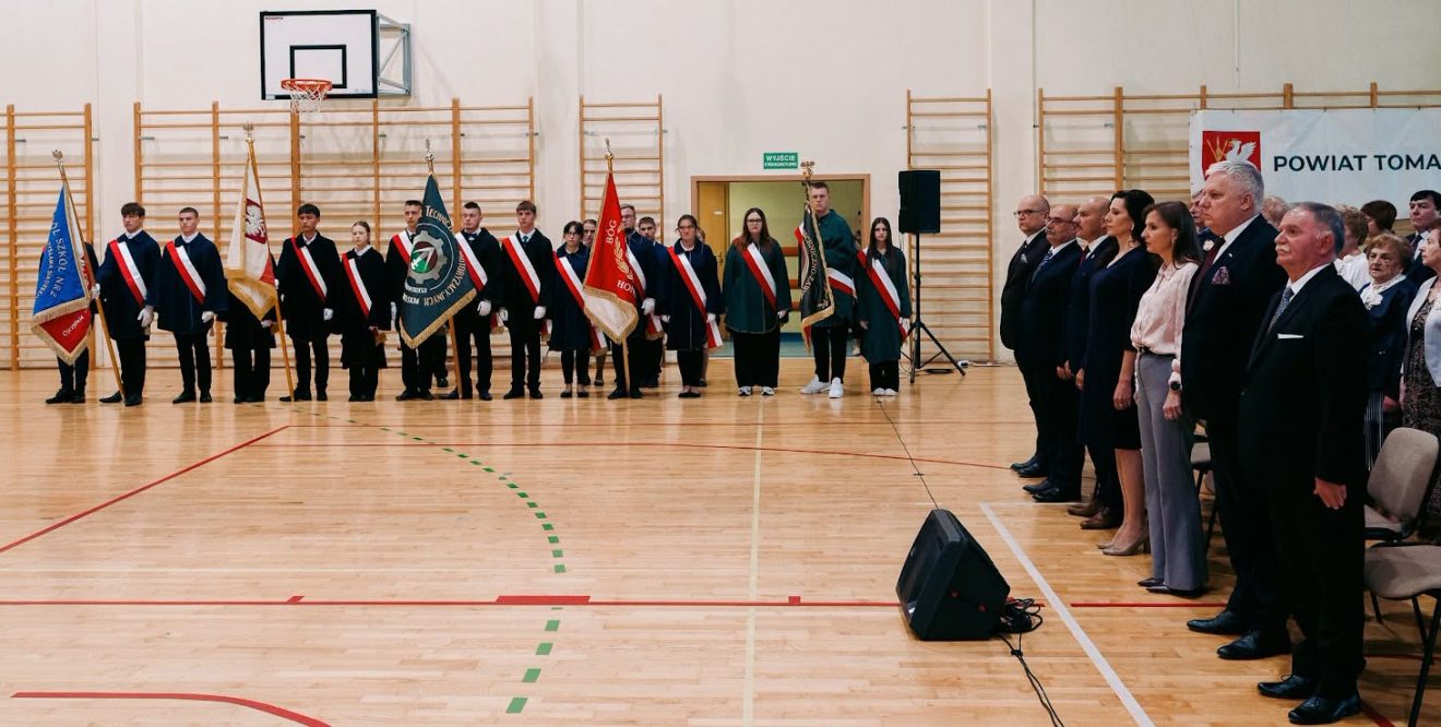 Poczet sztandarowy uczniów stoi w szeregu, a po prawej stronie zgromadzeni goście uczestniczą w uroczystości.
