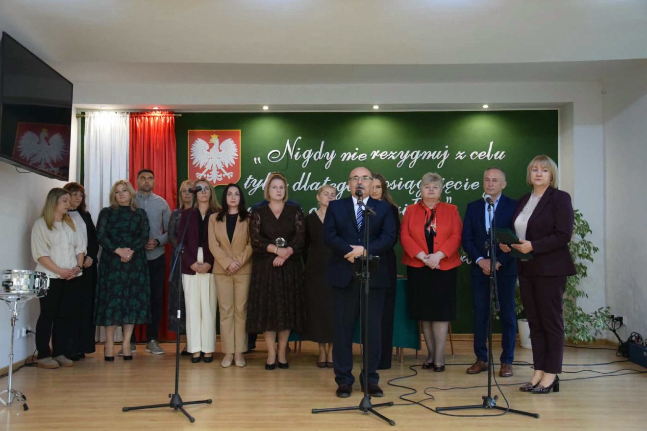 Przedstawiciele kadry szkolnej stoją na scenie podczas uroczystości, a Dariusz Czop Członek Zarządu Powiatu Tomaszowskiego przemawia do mikrofonu.