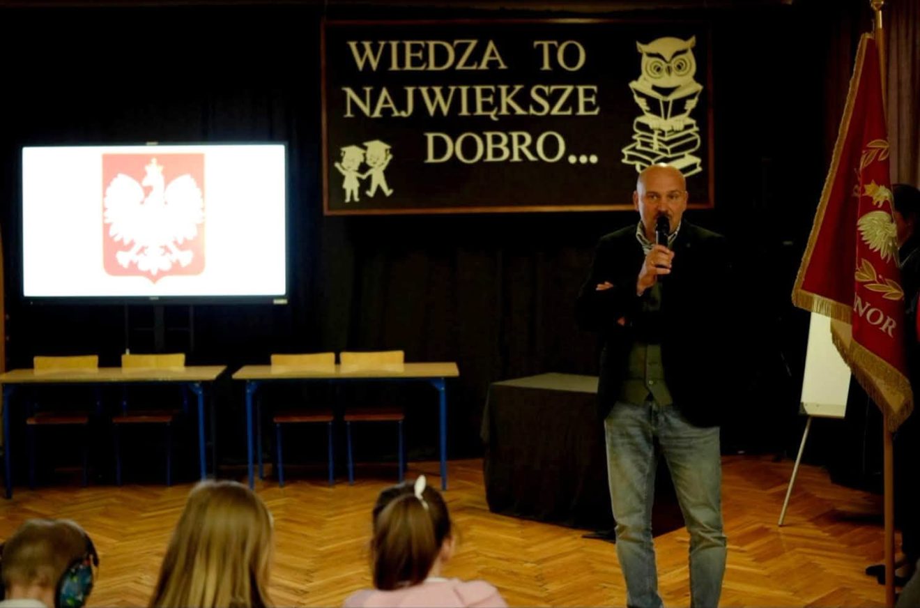 Wicestarosta Jarosław Korzeń przemawia do zgromadzonych na szkolnej sali. W tle widnieje napis „Wiedza to największe dobro” oraz godło Polski.